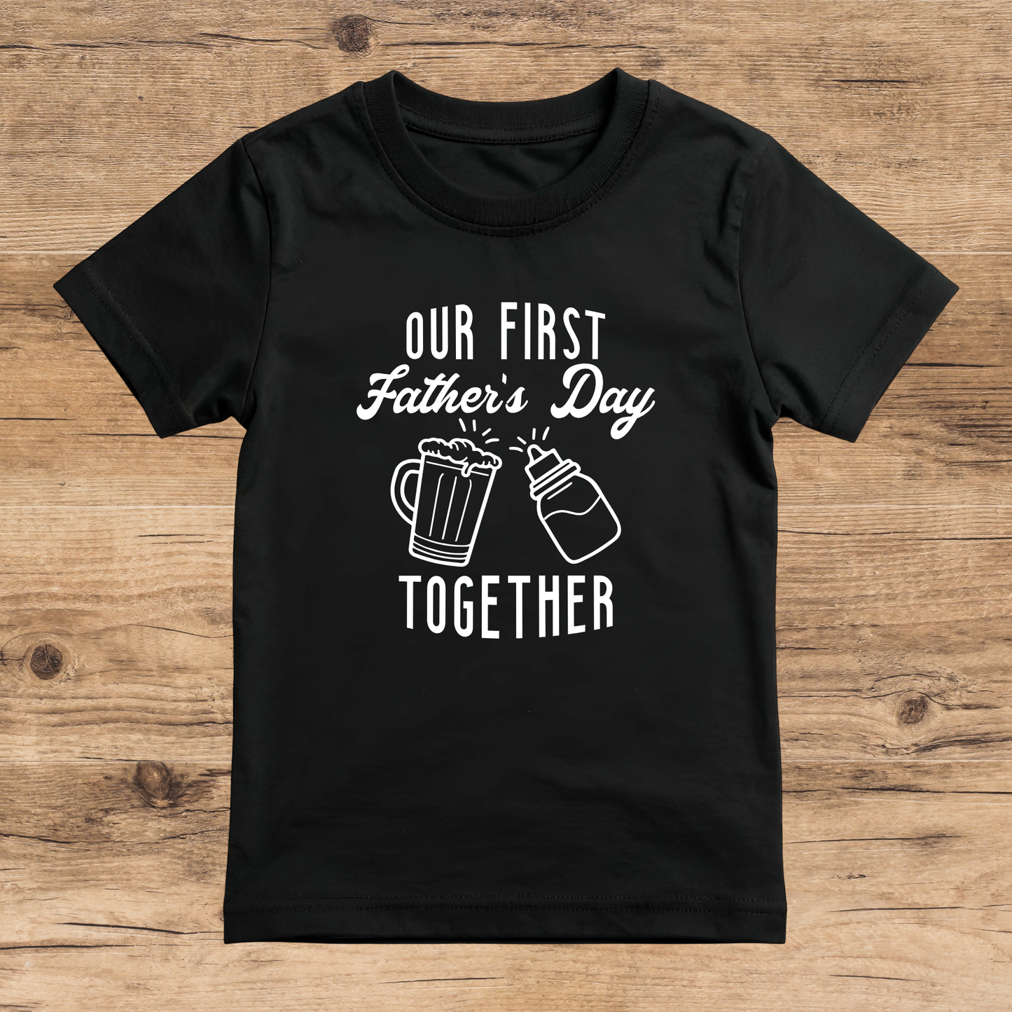 Black t-shirt with 'Our First Father's Day Together' text and beer mug graphics on a wooden surface