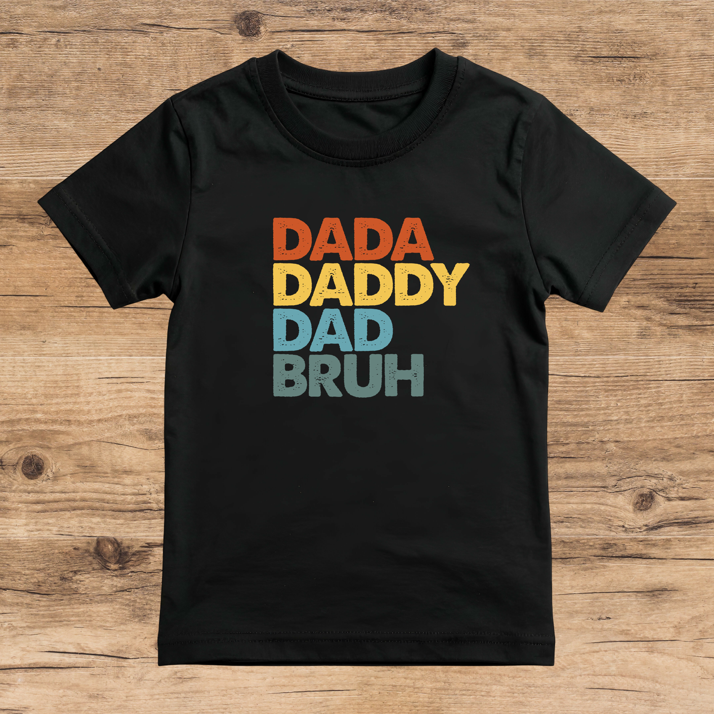 Black t-shirt with colorful text 'DADA DADDY DAD BRUH' on a wooden surface