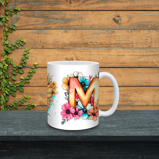 White mug with colorful letter 'M' design on a wooden surface with greenery.