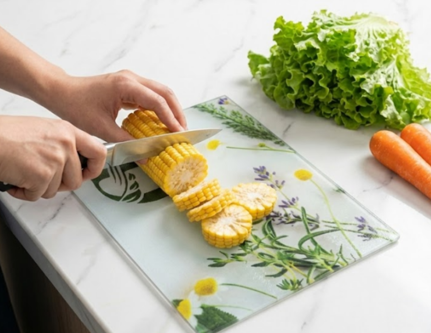 Person slicing corn on a decorative glass cutting board with vegetables in the background.