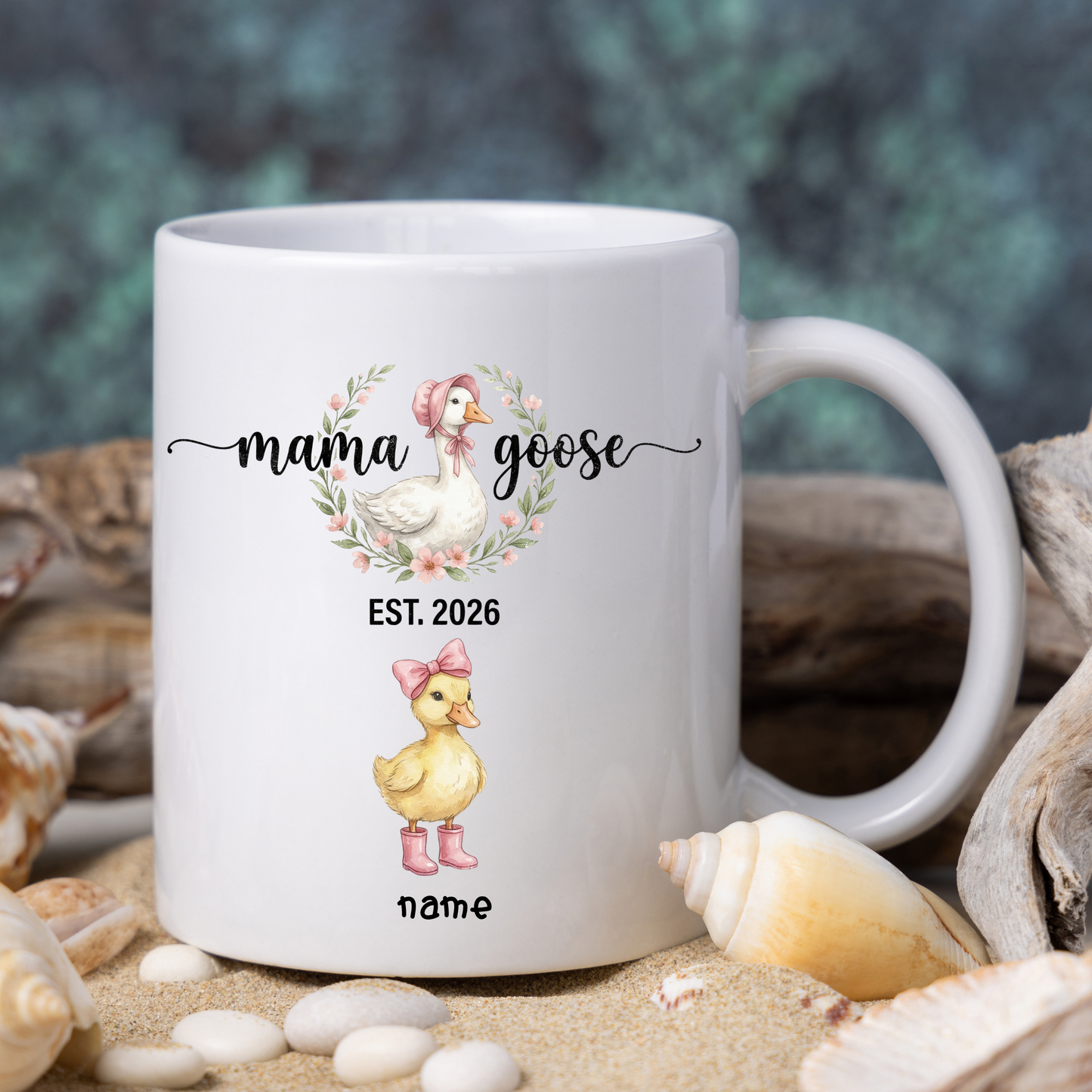 White mug with 'mama goose' design featuring a duck and text, surrounded by shells and sand.