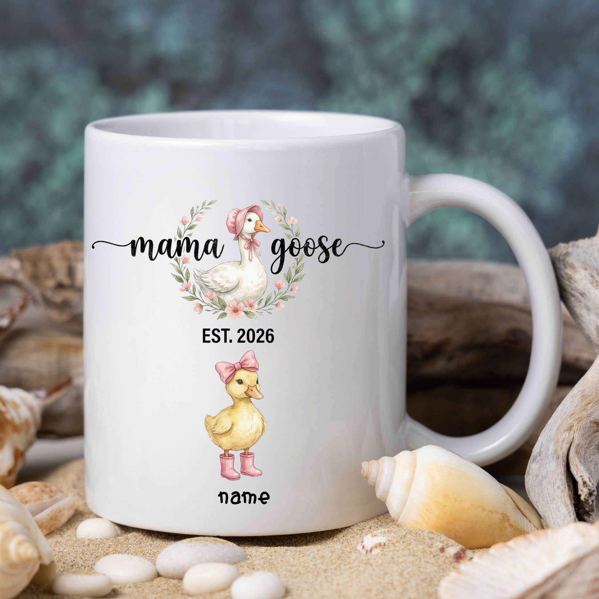 White mug with 'mama goose' design featuring a duck and text, surrounded by shells and sand.