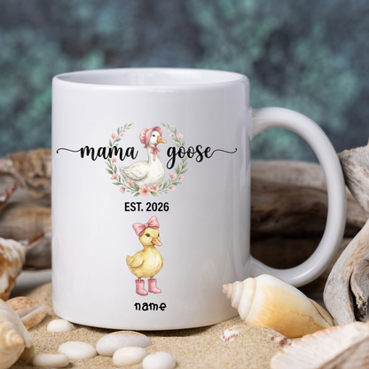 White mug with 'mama goose' design featuring a duck and text, surrounded by shells and sand.