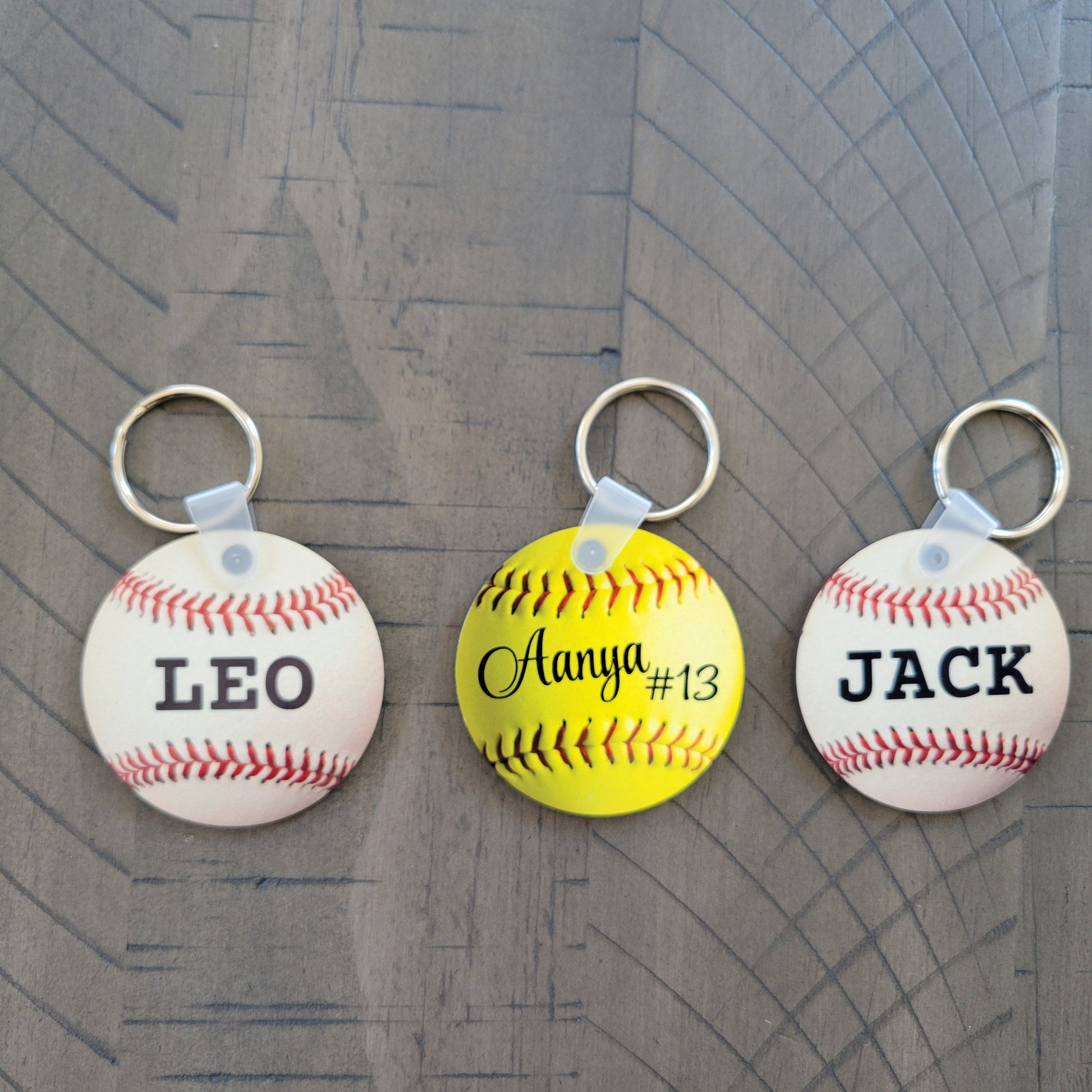 Three keychains shaped like baseballs and a softball with names engraved on them, placed on a wooden surface.