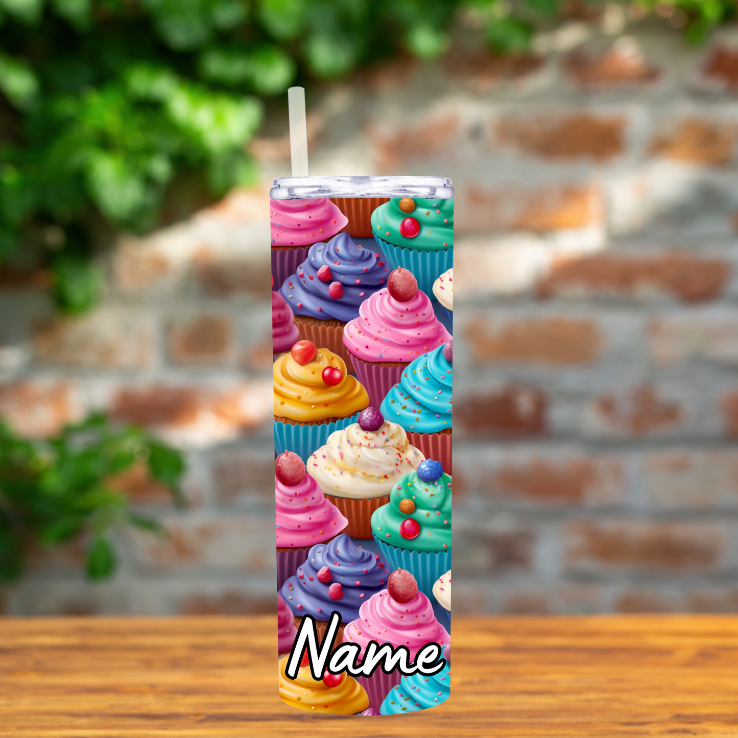 Tumbler with colorful cupcake design on a wooden surface with a blurred brick wall background