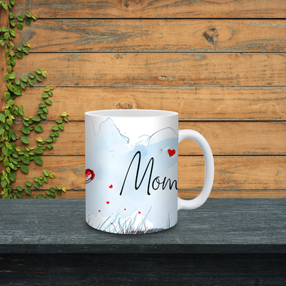White mug with 'Mom' printed on it, placed on a wooden surface with greenery.
