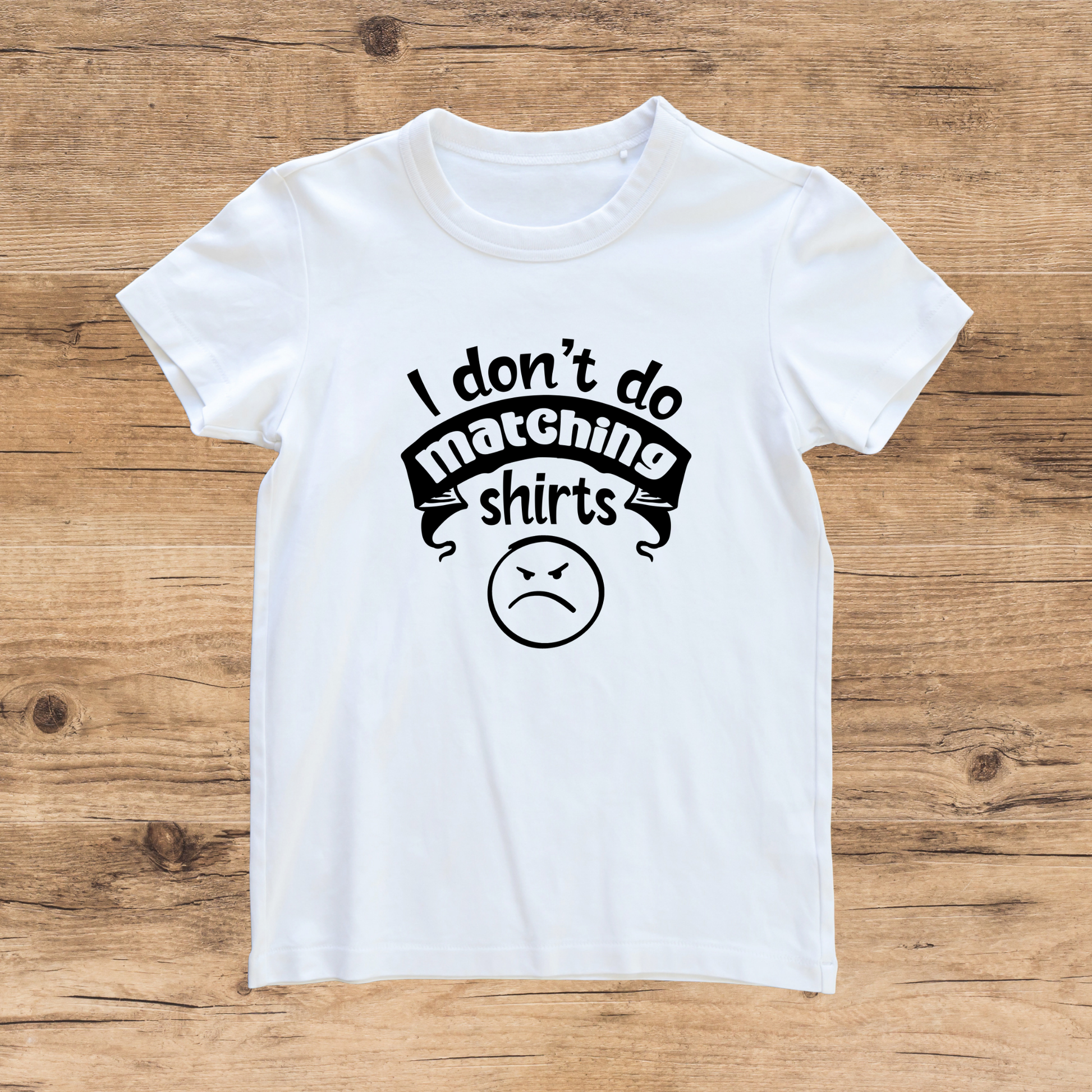 White t-shirt with black text and an angry face emoji on a wooden surface
