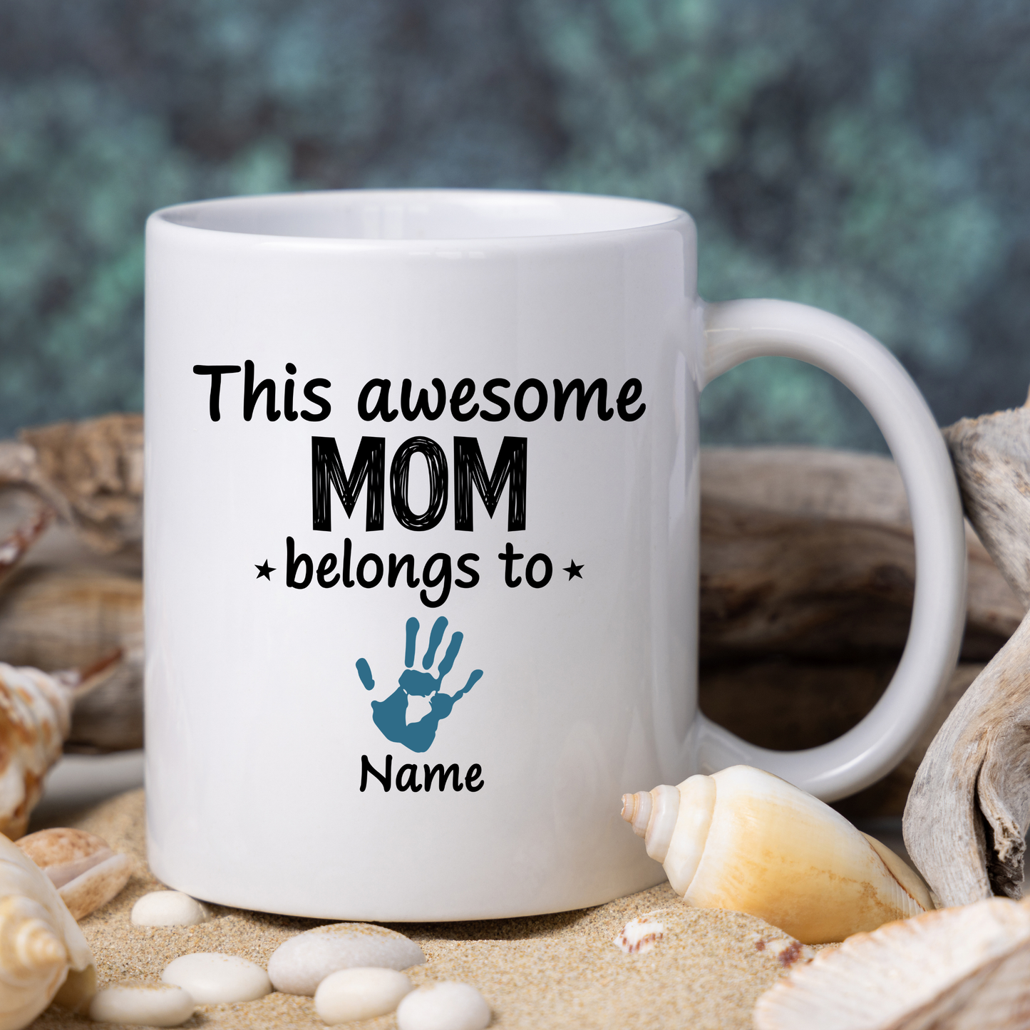White mug with 'This awesome mom belongs to' text and handprint design, surrounded by shells and sand.
