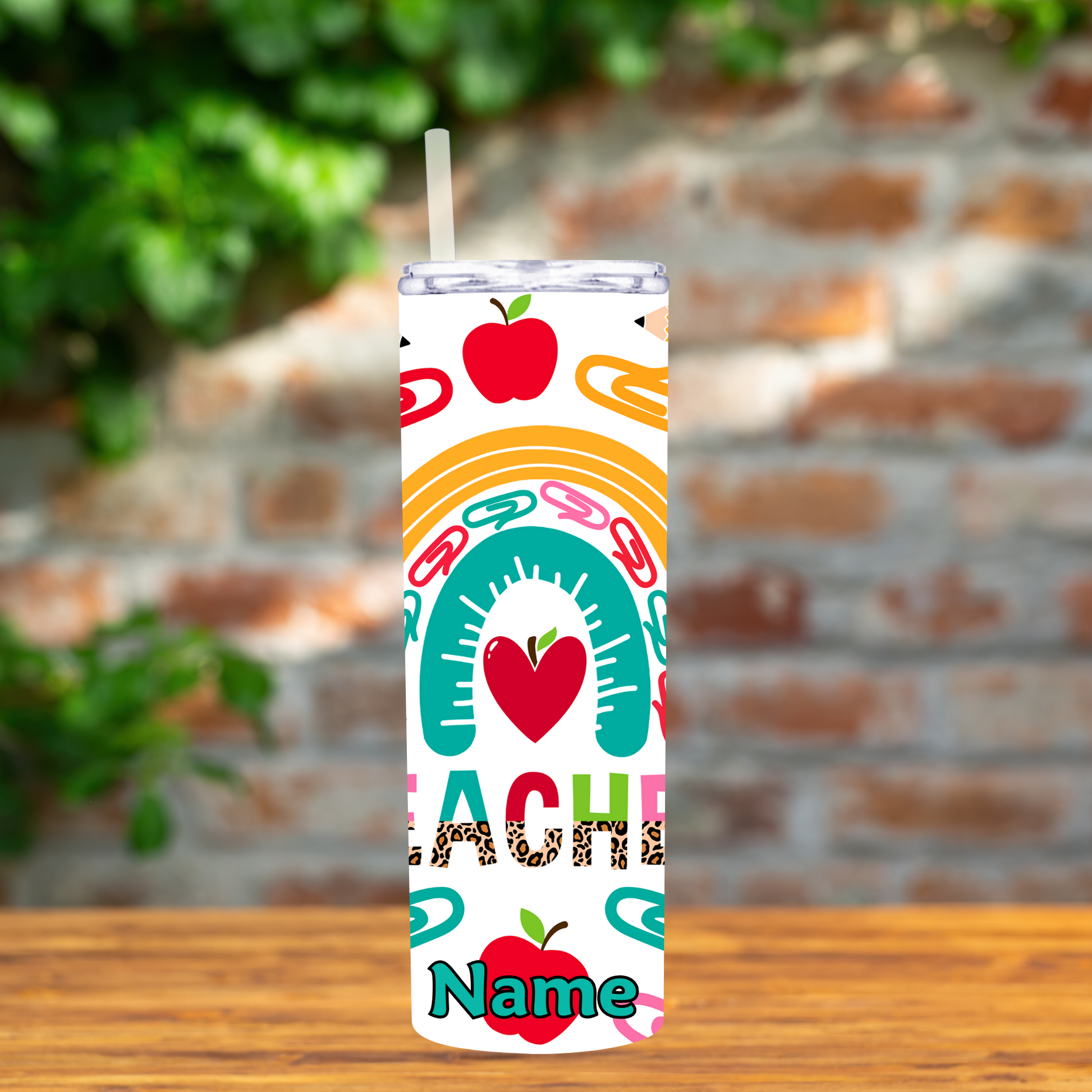 Tumbler with colorful design saying teacher featuring apples, hearts, and letters on a wooden surface with a blurred brick wall background.