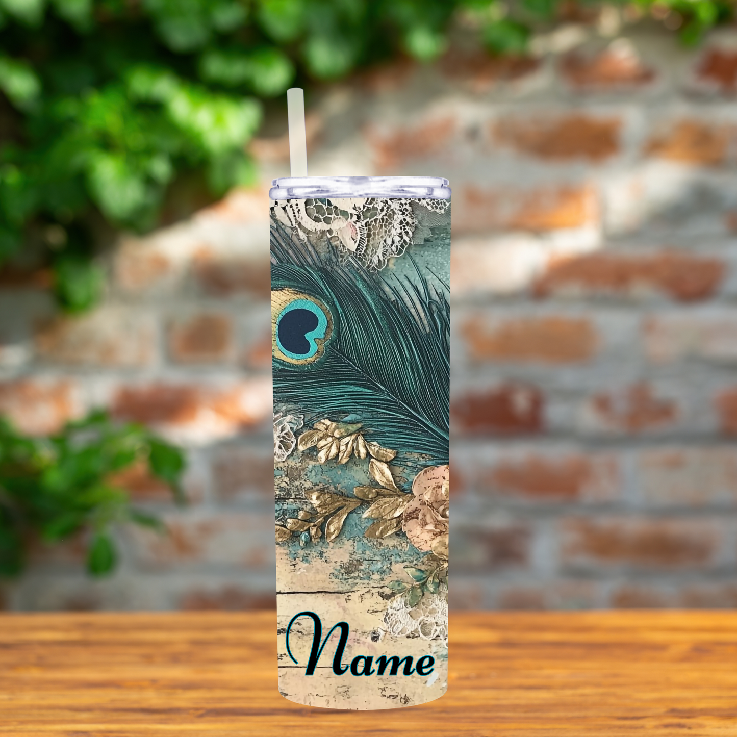 Tumbler with peacock feather and lace design and 'Name' text on a wooden surface with a blurred brick wall background.