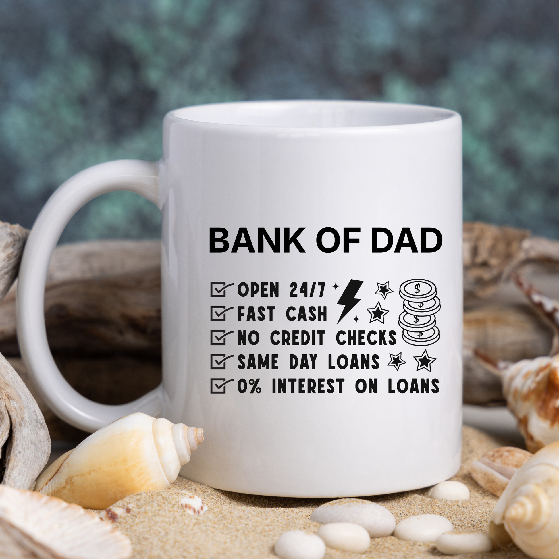 White mug with text 'Bank of Dad' surrounded by seashells on sand