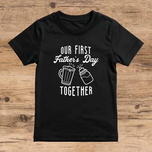 Black t-shirt with 'Our First Father's Day Together' text and beer mug graphics on a wooden surface