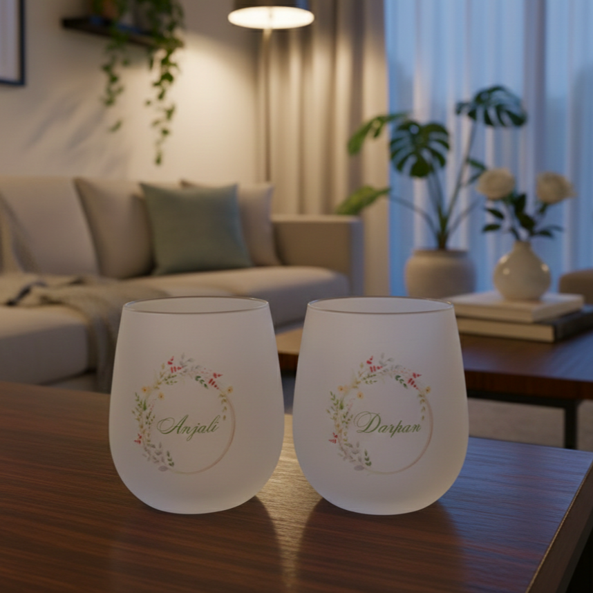 Two frosted wine glasses with floral designs and names on a wooden table in a cozy living room.