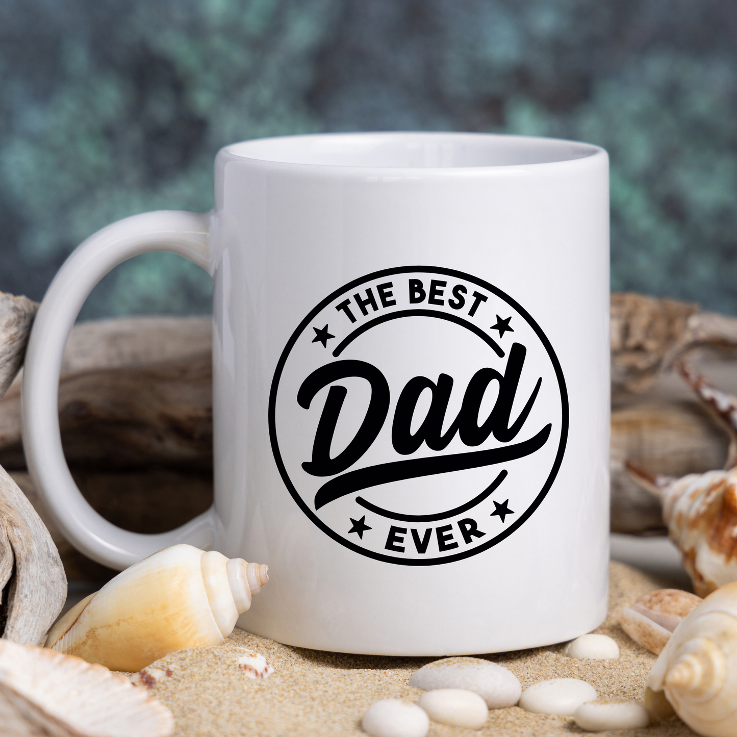 White mug with 'The Best Dad Ever' text on a beach setting with shells and sand.