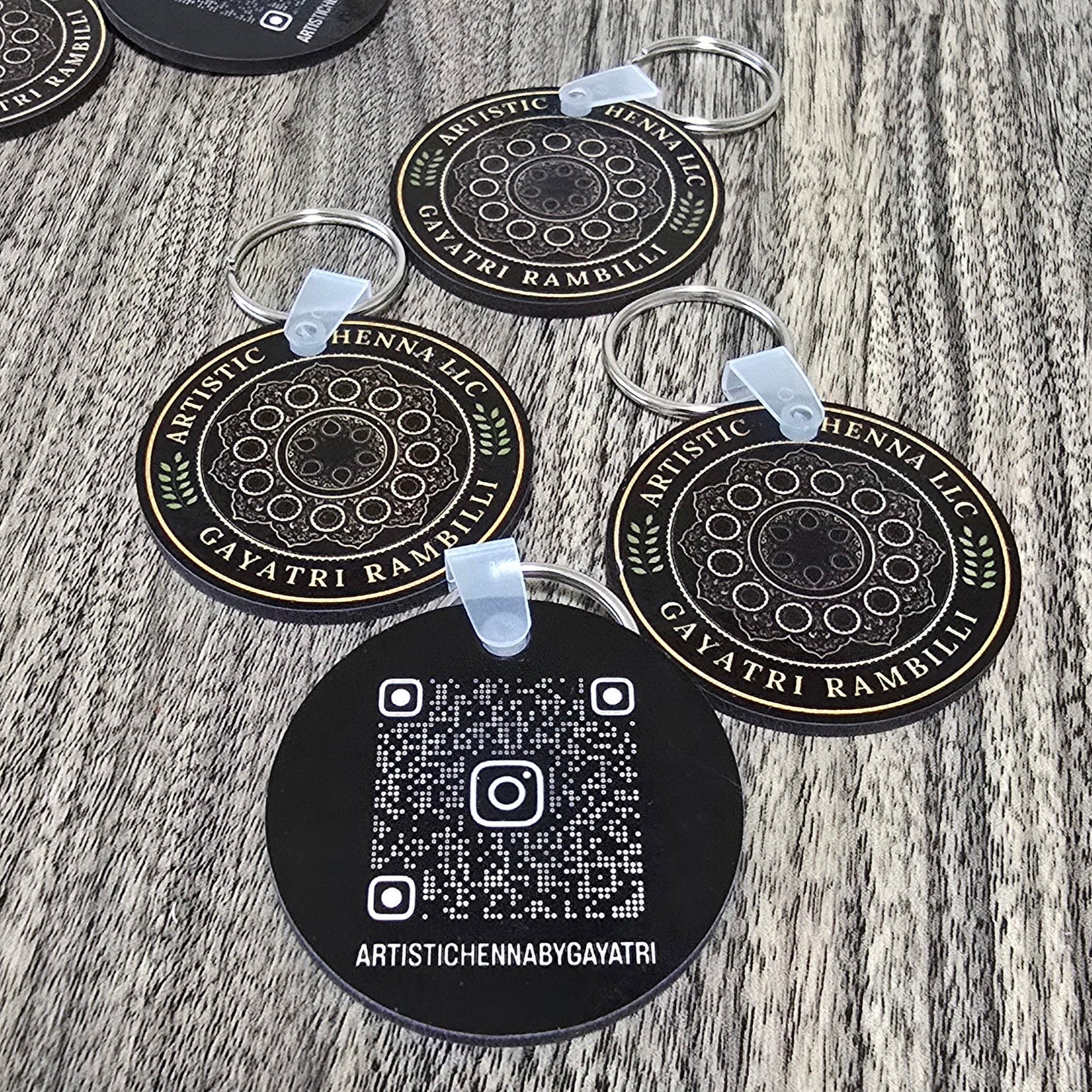 Several round black keychains with white printed mandala designs and QR codes are arranged on a gray wood-grain surface. The keychains have clear plastic loops attached.