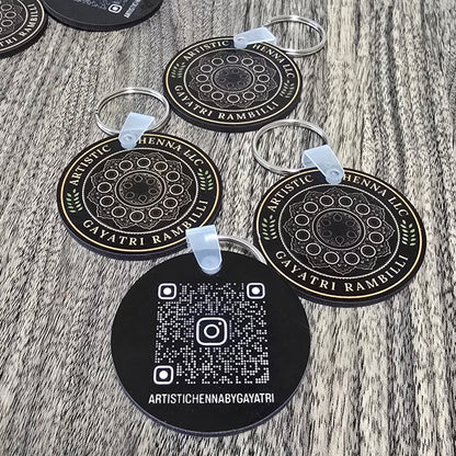 Several round black keychains with white printed mandala designs and QR codes are arranged on a gray wood-grain surface. The keychains have clear plastic loops attached.