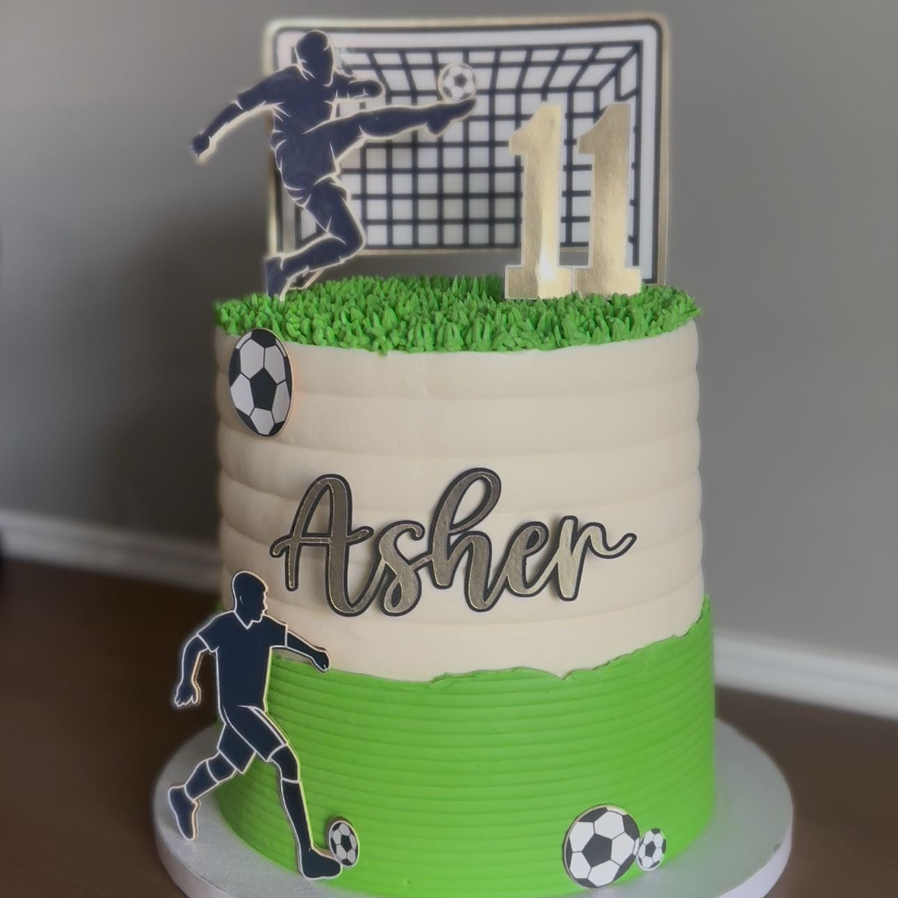 Soccer-themed birthday cake with player and goal topper, grassy base, and 'Asher' on a plain background.
