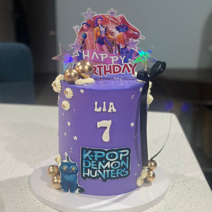 Purple birthday cake with 'Happy Birthday' and 'KPOP DEMON HUNTERS' text, featuring decorative elements on a white surface.