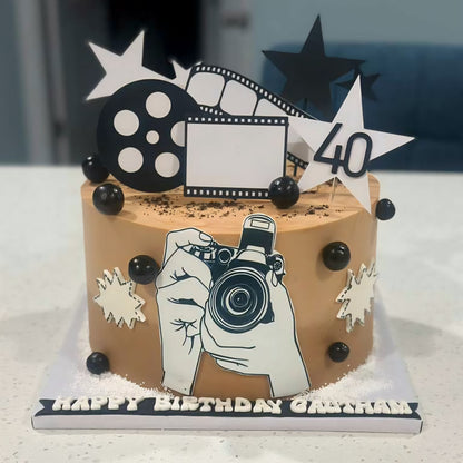 Birthday cake with camera-themed decorations and 'Happy Birthday' topper on a white surface.
