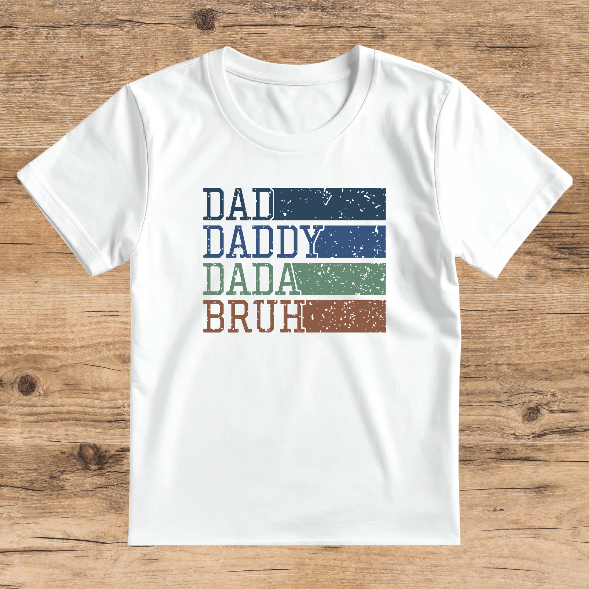 White t-shirt with 'DAD', 'DADDY', 'DADA', 'BRUH' text on a wooden surface
