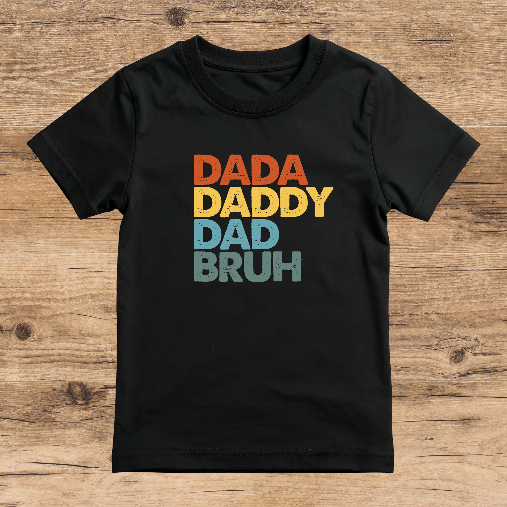 Black t-shirt with colorful text 'DADA DADDY DAD BRUH' on a wooden surface