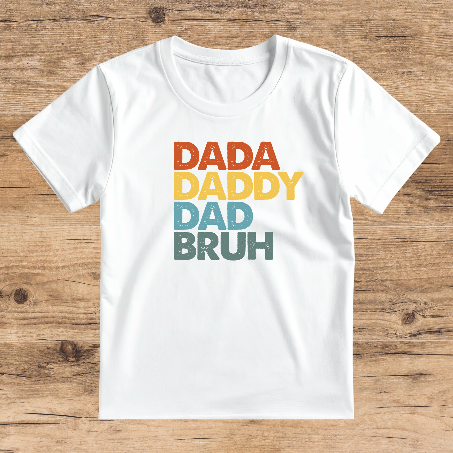 White t-shirt with colorful text 'DADA DADDY DAD BRUH' on a wooden surface
