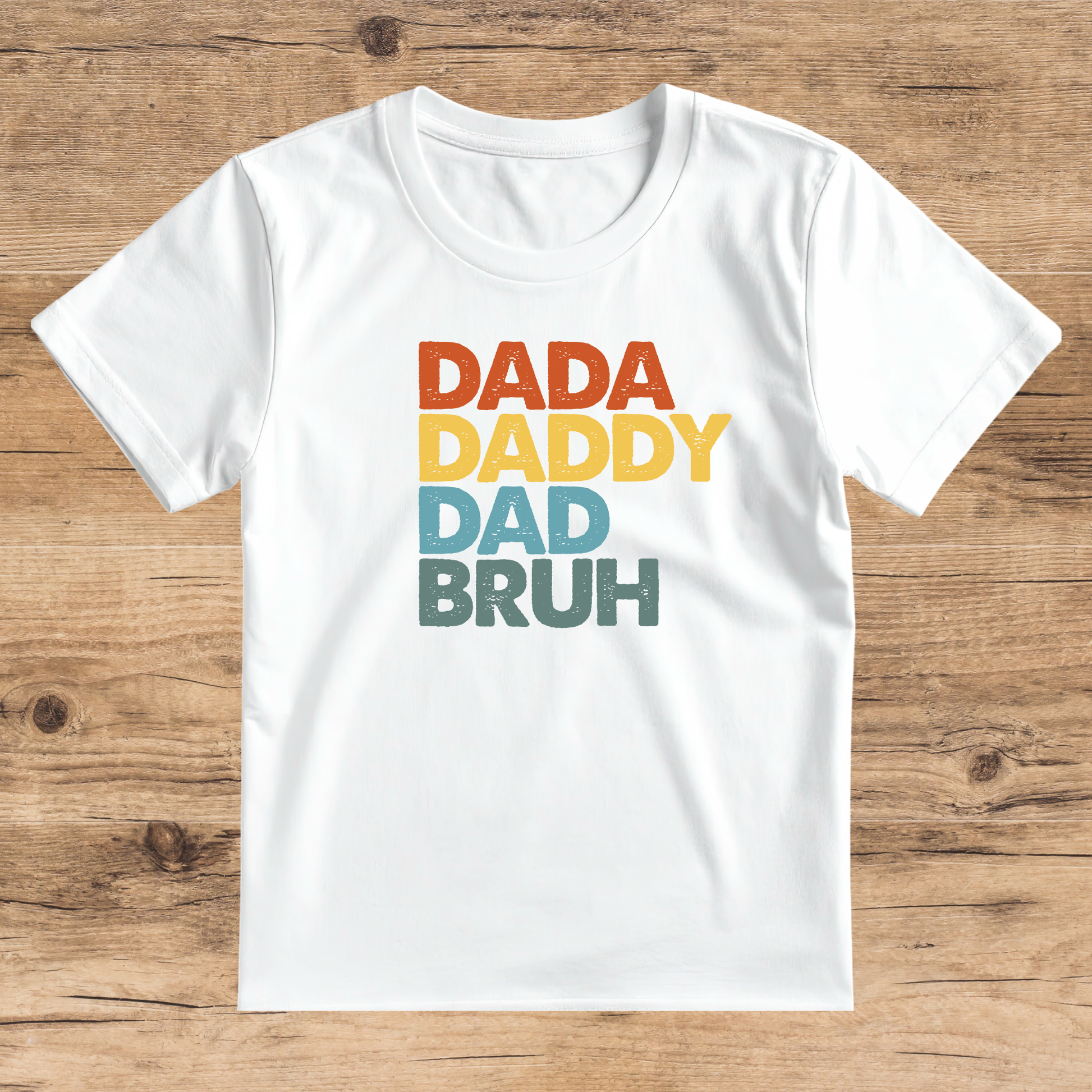 White t-shirt with colorful text 'DADA DADDY DAD BRUH' on a wooden surface