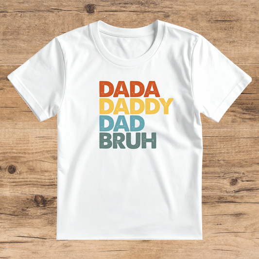 White t-shirt with colorful text 'DADA DADDY DAD BRUH' on a wooden surface