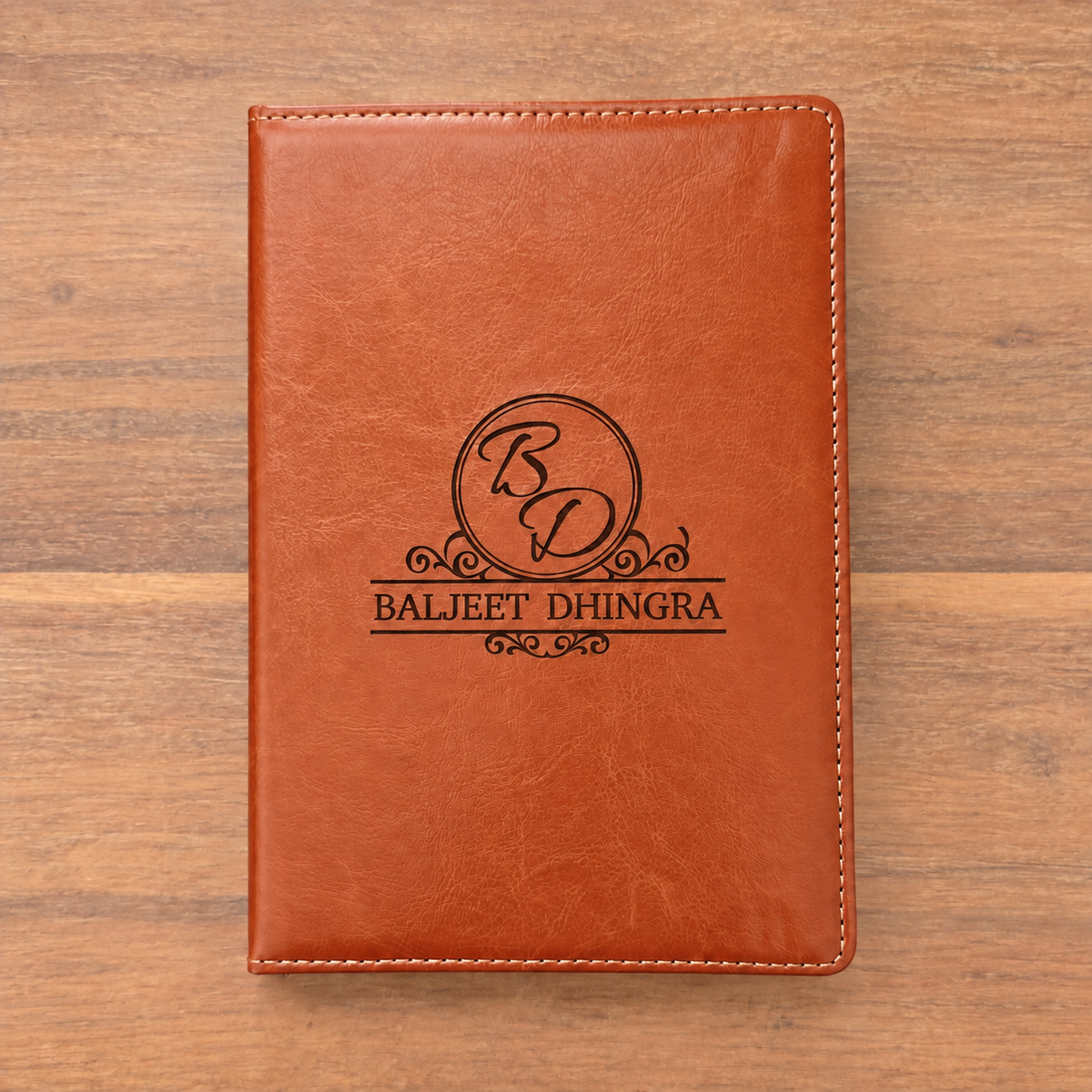 A brown faux-leather notebook cover with white stitching and a black engraved monogram logo reading 'BD' and 'BALJEET DHINGRA', shown flat on a wooden surface.