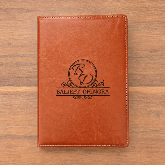 A brown faux-leather notebook cover with white stitching and a black engraved monogram logo reading 'BD' and 'BALJEET DHINGRA', shown flat on a wooden surface.