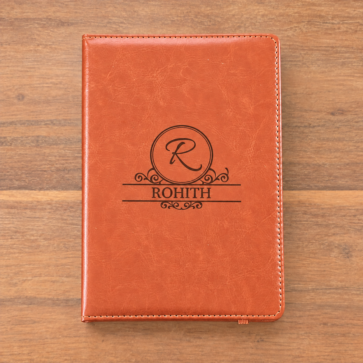 A personalized orange-brown faux-leather notebook cover with a black monogrammed design featuring the letter 'R' in a circle and the name 'ROHITH' below, displayed flat on a wooden surface.