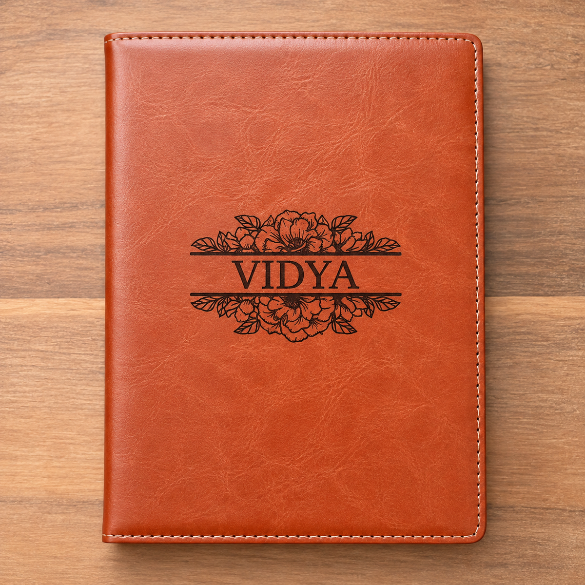A personalized brown faux-leather notebook cover with white stitching and a black floral engraving reading 'VIDYA', displayed flat on a wooden surface.