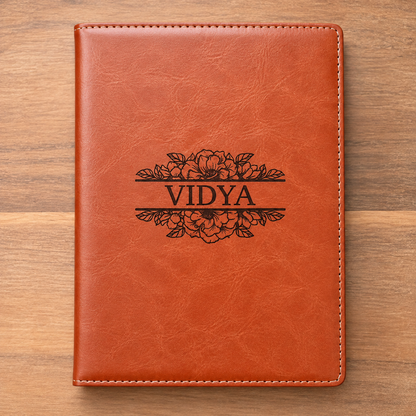 A personalized brown faux-leather notebook cover with white stitching and a black floral engraving reading 'VIDYA', displayed flat on a wooden surface.