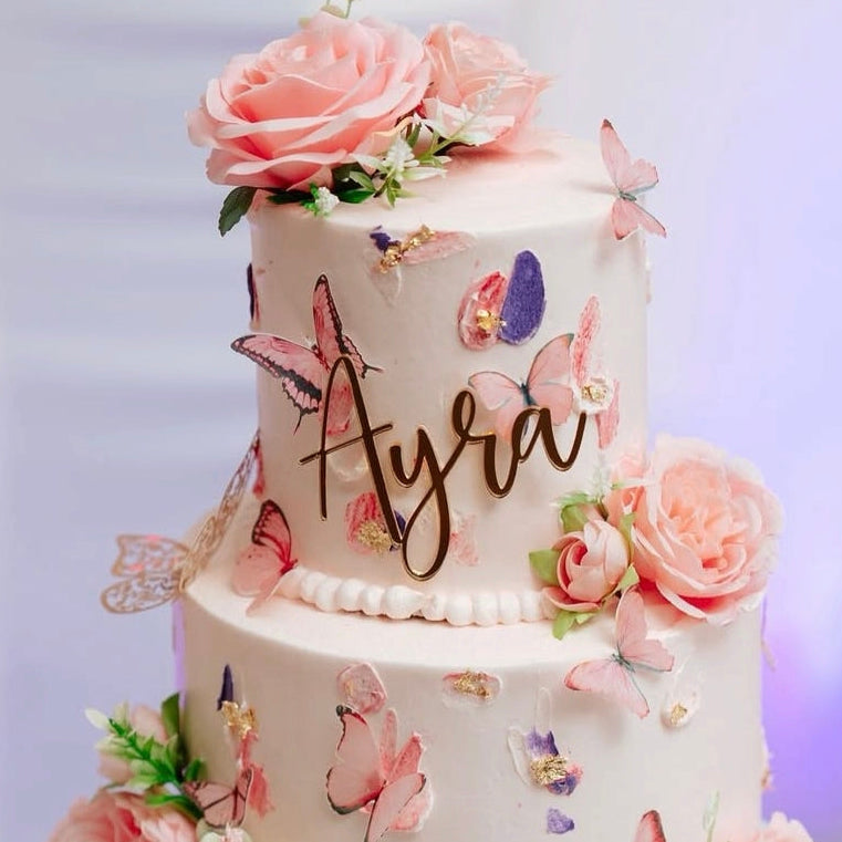 Two-tiered cake with floral decorations and butterfly designs, featuring the name 'Ayla'.