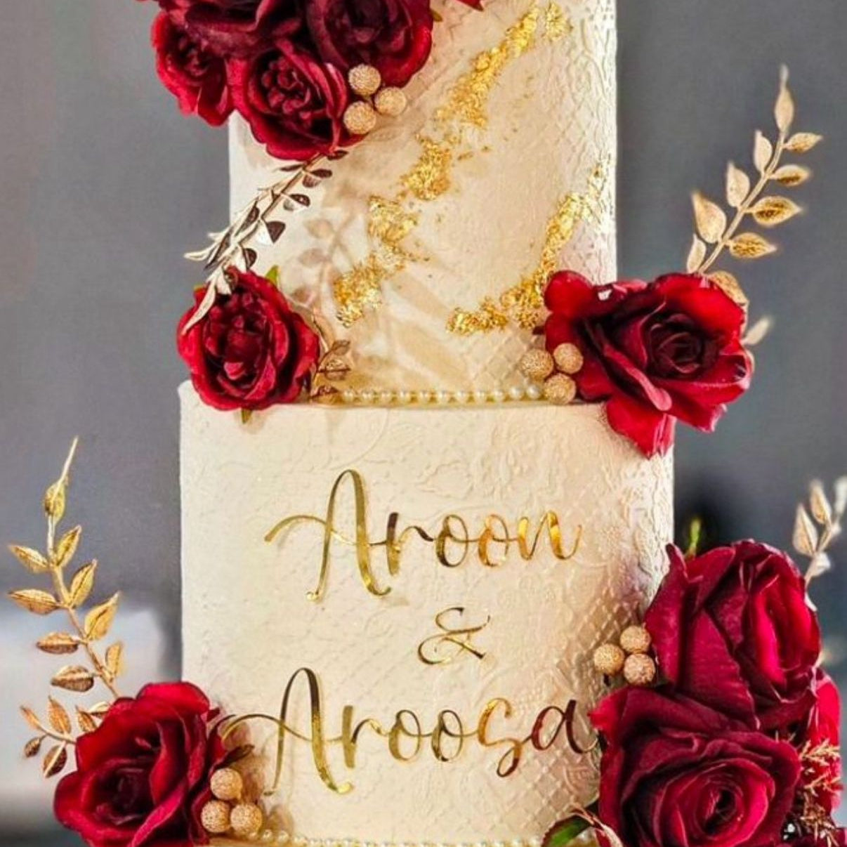 Three-tiered wedding cake decorated with red roses and gold accents, featuring the names 'Aroon & Aroosa'.